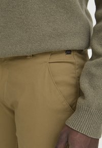 Olivgrüner Strickpullover kombiniert mit beigen Slim-Fit-Hosen. Der Pullover hat ein strukturiertes Muster; die Hose verfügt über Akzentnähte und ein kleines Logotag.