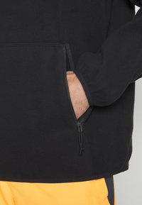 Pullover in pile nero con una tasca con zip e una superficie liscia. L'orlo inferiore è dritto, sovrapposto a pantaloni gialli.