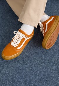 Brown suede sneakers with white side stripes, flat cream laces, and a gum rubber sole, worn with white socks and khaki pants.