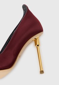 Burgundijas satīna augstpapēžu kurpe ar zelta metāla stiletto papēdi un gludu, izliekta dizaina virsmu, ar melnu iekšējo oderi.