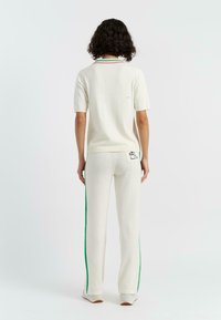 Tweedelige outfit van crème stof; kortebloemige top met een groene en rode kraag; bijpassende lange broek met groene zijstrepen en logo-detail.