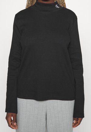 Zwarte top met lange mouwen en col met korte rits, met een kleine witte logo op de kraag, gecombineerd met lichtgrijze pantalon met fijne krijtstreep, gezien van nek tot heupen.