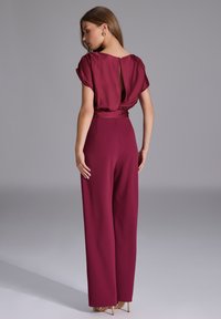 Burgundowa kombinezon z krótkimi bufkami, plisowanym tyłem i szerokimi nogawkami. Gładka tekstura materiału i widoczne złote akcenty.