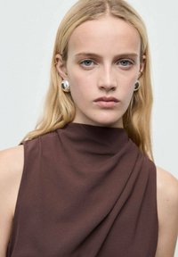 Model draagt een bruin, mouwloos kledingstuk met een hoge hals. Grote zilveren oorbellen zijn zichtbaar. Huid lijkt licht met steil blond haar.