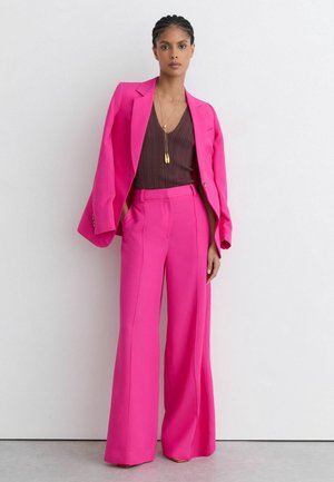 Frau mit geflochtenem Haar, die leuchtend pinke, weit geschnittene Hose und einen passenden Blazer über einem dunklen ärmellosen Oberteil trägt, steht vor einer schlichten Wand.