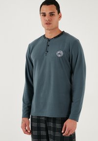 Garais piedurknes tumšzaļais krekls ar pogājamu henley dekoltē, uz kreisā krūšu puse ir apļveida logo, un apkakles iekšpusē ir ķekaru raksts.