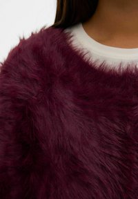 Bordo mākslīgās kažokādas apģērbs ar mīkstu, pūkainu tekstūru, apaļu kakla izgriezumu un slāņotu dizainu uz gaišas pamatnes.