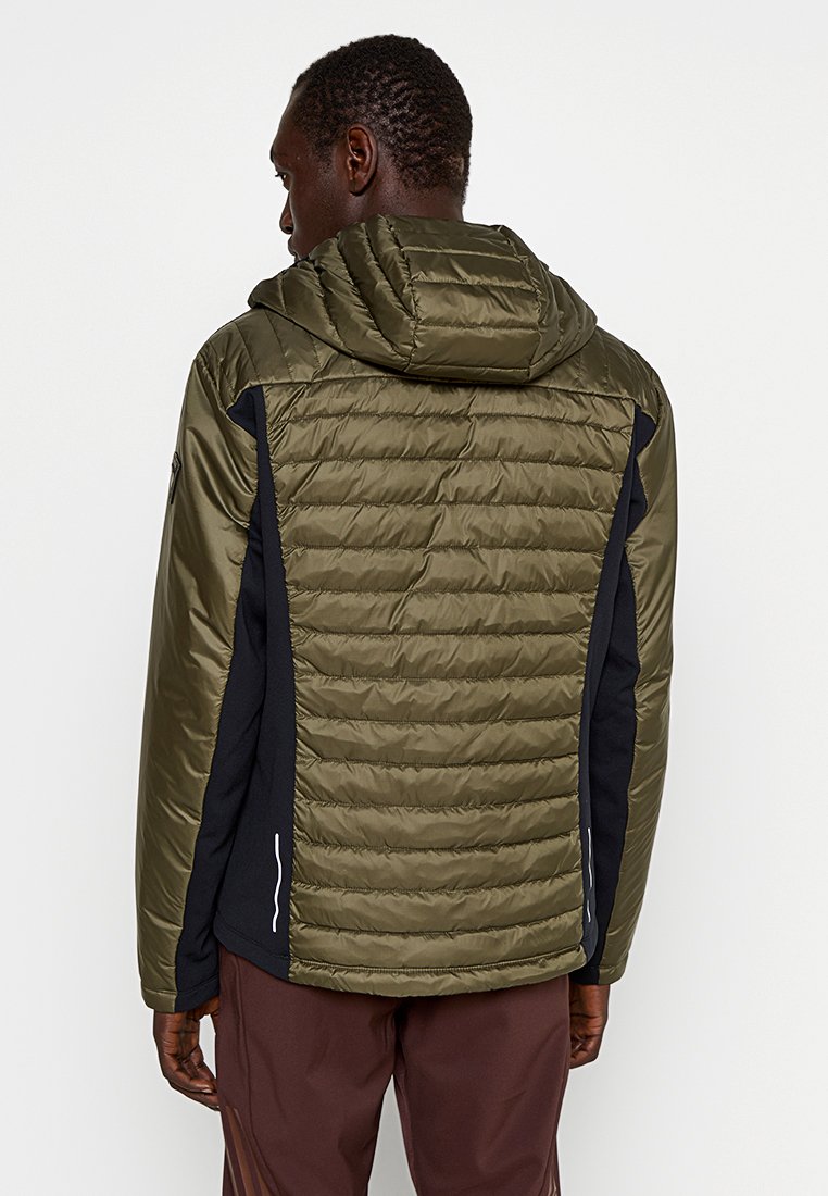 Man met zijn rug naar de camera, gekleed in een olijfgroene gewatteerde jas met capuchon en zwarte zijpanelen, gecombineerd met bruine broeken.