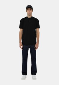 Polo shirt nera con colletto classico, maniche corte, abbinata a jeans blu scuro e sneaker bianche. Il modello indossa anche un berretto grigio.