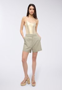 Body con dorato metallizzato, pantaloni corti verdi chiari a vita alta e sandali beige con plateau e cinturini incrociati. Texture liscia e design aderente.