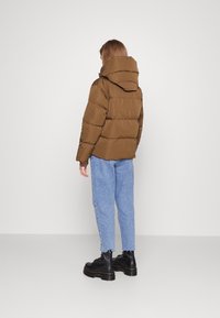 Chaqueta acolchada marrón con capucha, con un diseño de rombos. Combinada con jeans de color azul claro y botas negras gruesas. Silueta simple y estilizada.