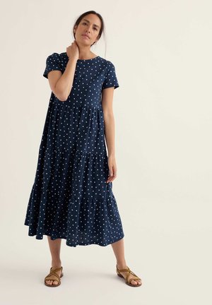 Vrouw met donker haar in een marineblauwe midi-jurk met korte mouwen en polka dots, en bruine sandalen met bandjes, staand tegen een effen lichte achtergrond.