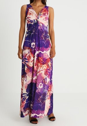 Vrouw draagt mouwloze maxi-jurk met grote bloemenprint in paars, rood en roze en knoopdetail op de borst, gecombineerd met zwarte open-toe hakken.