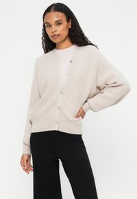 Cardigan acanalado de color beige claro con cuello en V, cierre de botones y mangas largas. Combinado con pantalones negros, presentado sobre un fondo blanco.