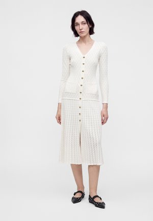Femme aux cheveux courts et foncés portant une robe blanche texturée boutonnée et des ballerines Mary Jane noires cloutées, debout devant un fond uni.