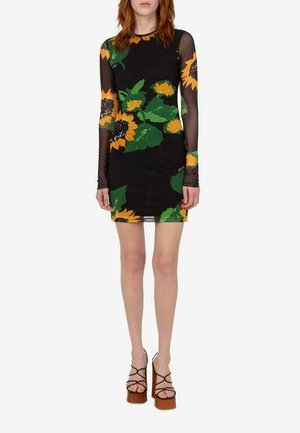 Mujer que lleva un vestido mini negro transparente de manga larga con grandes patrones de girasoles y hojas verdes y sandalias marrones con plataforma.