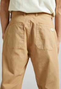 Pantalones de algodón en tono beige que cuentan con un corte relajado, dos bolsillos traseros y una pequeña etiqueta tejida en el bolsillo derecho. Textura suave con un acabado mate.