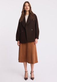 Brązowy płaszcz dwurzędowy z mieszanki wełny, luźny krój, noszony na beżowej teksturowanej bluzce i brązowej spódnicy w kształcie litery A, zestawiony z czarno-beżowymi szpilkami w cętki.