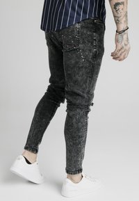 Jeans ajustados negros con un lavado desgastado, con detalles de salpicaduras en rojo y blanco, combinados con zapatillas blancas.