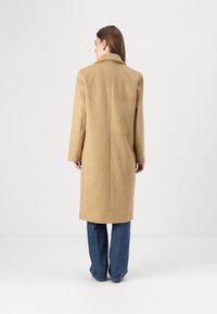 Cappotto di lana beige con colletto a taglio, maniche lunghe e silhouette dritta, indossato sopra jeans a gamba larga blu. Vista posteriore, texture liscia.