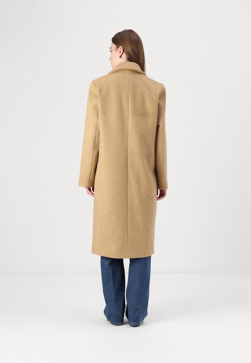 Beżowy wełniany płaszcz z klapami, długimi rękawami i prostym krojem, noszony na szerokich niebieskich dżinsach. Widok z tyłu, gładka tekstura.