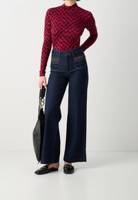 Jeans in denim blu scuro a vita alta con gambe larghi e tasche decorative. Maglietta rossa con motivo a maniche lunghe. Borsa e scarpe in pelle nera.