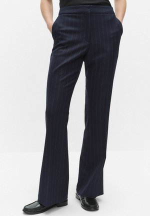 Person som bär marinblå skräddarsydda byxor med kritstrecksrand, med händerna i fickorna och svarta loafers mot en enfärgad bakgrund.