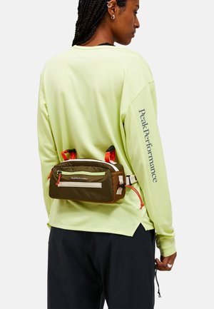 Bruin en groen heuptasje met oranje accenten en witte ritssluiting; bevestigd aan de rug van een model, gedragen over een limegroene lange mouw shirt.