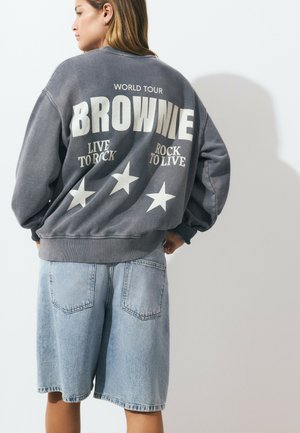 Woman wearing a gray oversized sweatshirt with "BROWNIE" text and stars, paired with light blue denim shorts, standing against a white wall.