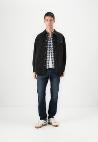 Chaqueta de pana negra sobre una camisa de cuadros azul y blanca, combinada con jeans oscuros y zapatillas blancas con suelas marrones.