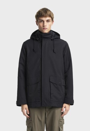 Chaqueta negra impermeable con capucha, cierre de cremallera, cuerdas ajustables y dos bolsillos frontales con solapa. Textura lisa y mate.