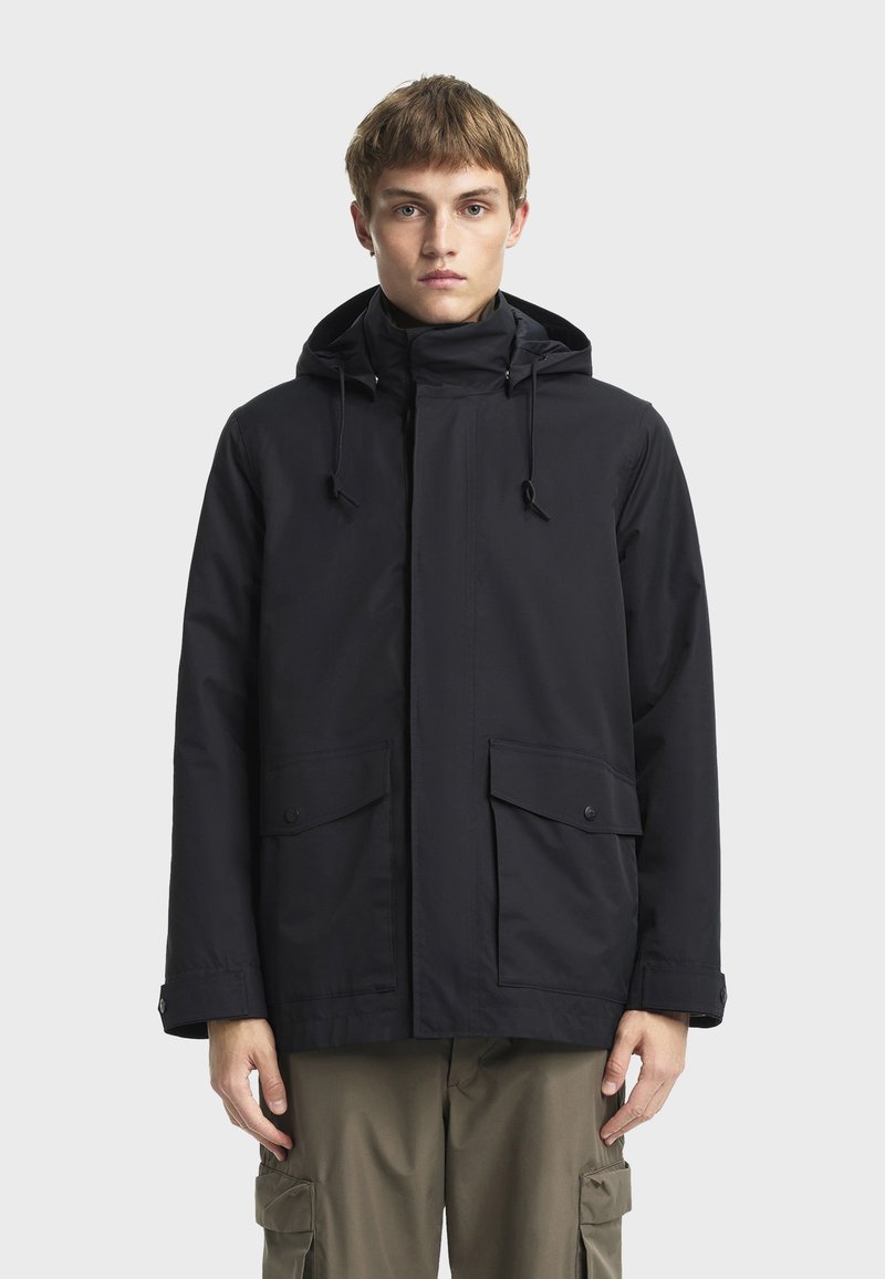 Crna vodootporna jakna s kapuljačom, zatvaranjem na zip, podesivim vezicama i dva prednja džepa s preklopom. Glatka, mat tekstura.