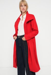 Cappotto rosso in lana con colletto alto, caratterizzato da una fila di bottoni e spacco laterale, indossato sopra una camicia chiara e jeans scuri.