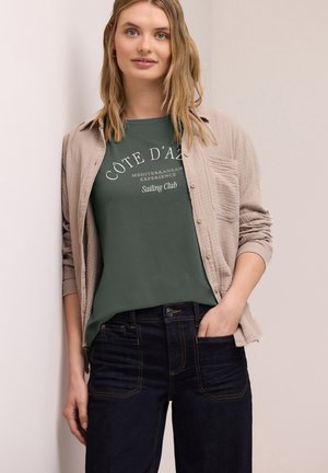 Joven con cabello rubio hasta los hombros que lleva una camiseta verde con el texto "Côte d'Azur Sailing Club" bajo una camisa beige abotonada y jeans oscuros.