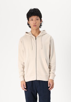 Hooded zip-up sweatshirt in light beige, made of soft fabric, featuring a front zip closure and two side pockets, with ribbed cuffs and hem.