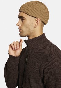 Brun strikket sweater med høj krave og lynlås, parret med en beige ribbet hue, båret af en model i profil mod en hvid baggrund.