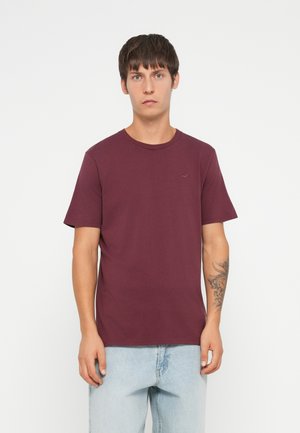 T-shirt corto color bordeaux in morbido cotone con collo rotondo e un piccolo logo sul petto, abbinato a jeans azzurri chiari.