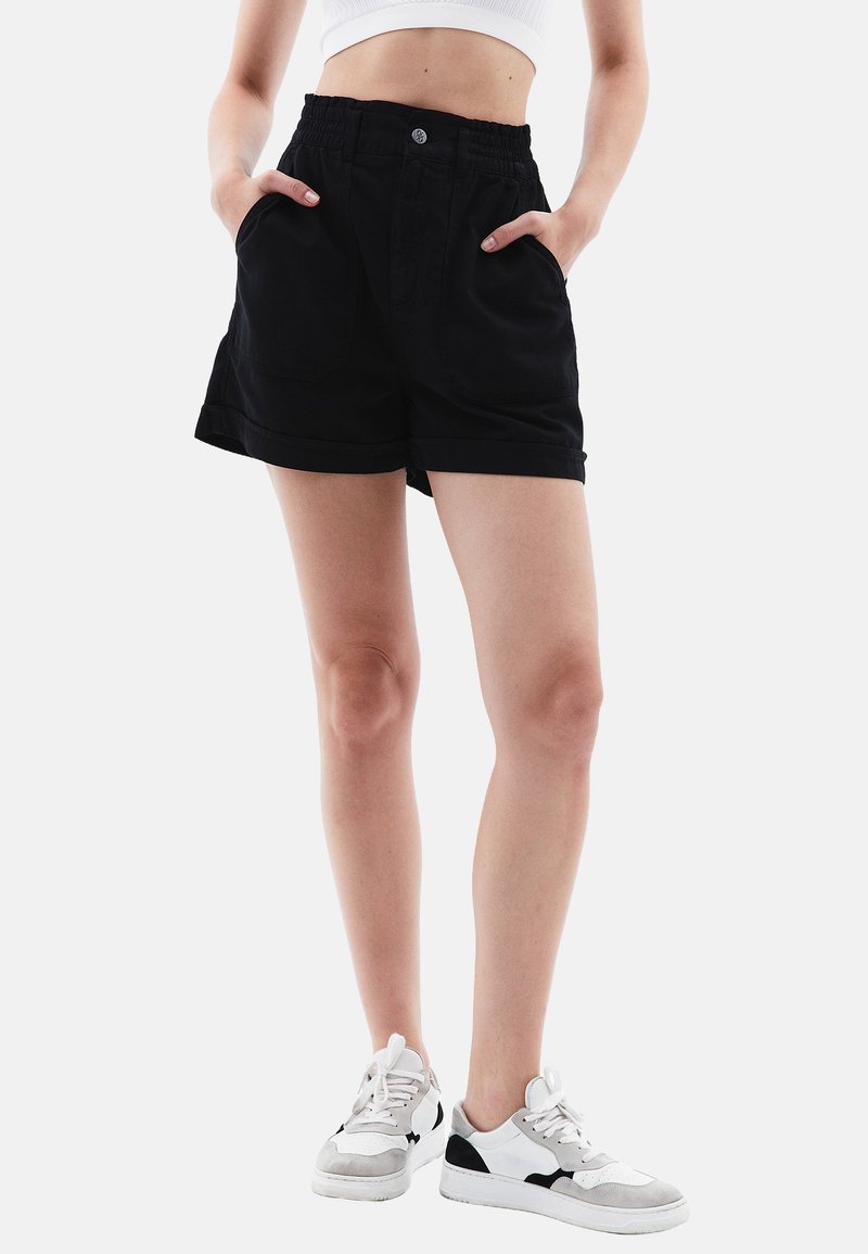 Pantaloni corti neri in cotone con vita elasticizzata, due tasche laterali e orlo arrotolato. Abbinati a sneakers bianche e grigie.
