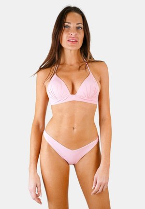 Femme aux longs cheveux bruns portant un bikini rose clair, debout devant un fond clair uni, tournée vers l'avant.