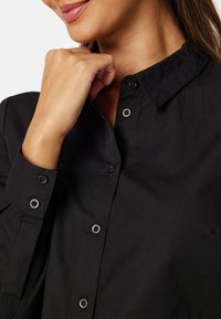 Camicia nera con colletto, realizzata in tessuto liscio, con bottoni grigi e una leggera texture stropicciata nella zona del colletto.