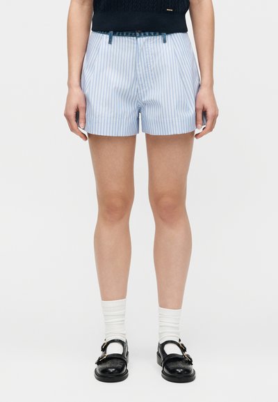 Άτομο που φοράει σορτς με κάθετες ριγέ σε ανοιχτό μπλε και λευκό χρώμα, λευκές κάλτσες και μαύρα παπούτσια με αγκράφα, στέκεται μπροστά σε απλό φόντο.