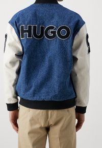 Kurtka bombowiec z denimu z niebieskim korpusem, kremowymi rękawami, prążkowanym kołnierzem oraz dużym czarnym napisem „HUGO” na plecach.
