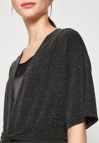 Svart wrap-top med korta ärmar, texturerad tyg med tunna glittrande trådar och djup V-ringning. Slät foder under.