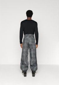 Moški stoji obrnjeno in nosi črn dolg rokav, sivo kariraste široke hlače z elastičnimi manšeti in črne škornje pred enobarvno ozadje.