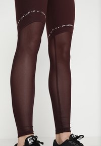 Leggings sportivi color bordeaux con un pannello semi-trasparente, dettagli cuciti e testo motivazionale bianco lungo l'elastico in vita.