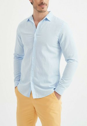 Homme portant une chemise à manches longues bleu clair boutonnée et un pantalon jaune moutarde, mains dans les poches, fond neutre.