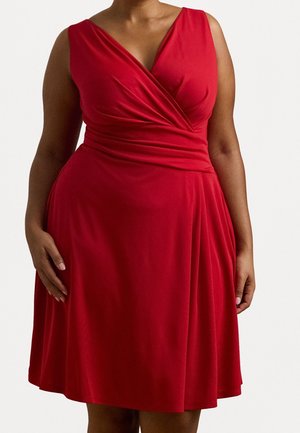 Femme portant une robe portefeuille rouge sans manches avec un col en V et une taille froncée, montrée du cou aux genoux sur un fond uni.