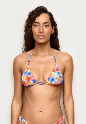 Femme aux longs cheveux bouclés portant un haut de bikini fleuri coloré avec des bretelles fines et des perles décoratives, debout devant un fond uni.