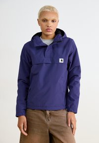 Anorak violet avec un col zippé mi-haut, capuche ajustable et poches latérales. Présente un patch logo à l'avant. Porté avec un pantalon marron.