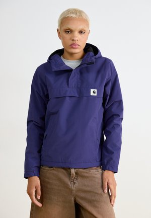 Paarse anorak jas met een halve rits, verstelbare capuchon en zijzakken. Heeft een logo patch aan de voorkant. Draagt met een paarse broek.